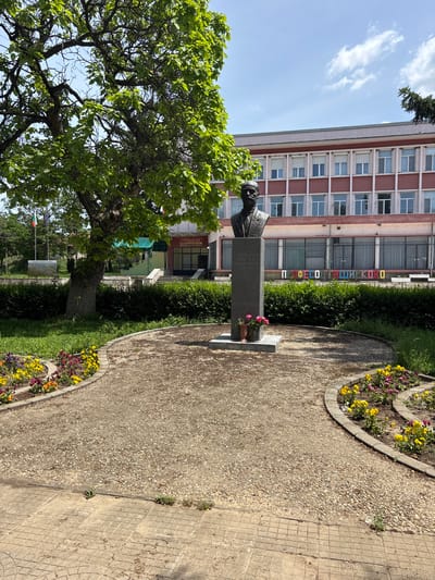 The image shows a statue of a man, likely a bust, situated in a park or garden in Profesor Ishirkovo, Bulgaria. The statue is placed on a stone pedestal. In front of it, pink flowers are set on the pedestal. A tree with green foliage is beside the statue on the left. A walkway curves around the statue, lined with flowerbeds containing yellow and purple flowers. The backdrop features a multi-story building with a red and white facade and windows. Letters spelling out "ПРОФЕСОР ИШИРКОВО" (Profesor Ishirkovo) are displayed on the building's exterior. Further behind the statue and building, there are other trees and vegetation. A flag (possibly the Bulgarian flag) is visible in the background.