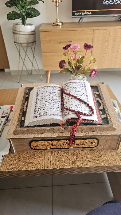 The scene depicts an indoor domestic environment, identified as a private residence in Sousse, Tunisia. In the immediate foreground, a light-brown, textured wooden coffee table is present. Centered on this table is an open Holy Quran, positioned on a carved wooden rehal (Quran stand). The Quran displays white pages with black Arabic script and red decorative borders. A red string of prayer beads (misbaha) with tassels is draped across the open pages. The front face of the rehal features a rectangular plaque with engraved Arabic script.

In the mid-ground, a light-colored wooden console table or low cabinet, equipped with two drawers and two cabinet doors, is situated against a light-colored wall. On the surface of this console, from left to right, are a golden, ornate candlestick base, and a clear vase containing a bouquet of artificial flowers in shades of pink and maroon, accompanied by green foliage. Directly behind these items, a flat-screen television is visible, displaying Arabic text which includes the names "عبد العزيز" (Abd al-Aziz) and "محرم" (Muharram). To the left of the console, a tall green potted plant in a white, possibly marble-patterned pot rests on a slender, gold-colored metal stand. The floor visible beneath the furniture is composed of light-colored, square tiles. No individuals or active interactions are present in the image.