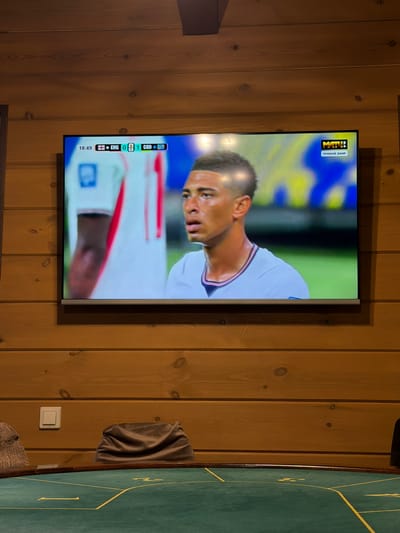 A television screen displays a soccer match, with a close-up of a player on the screen. The player is wearing a white jersey with red trim. The screen shows the score as ENG 0, GBR 1, and the time as 18:49. A "MATCH!" logo and "Прямой эфир" (Live broadcast) text are visible in the top left corner of the screen. The television is mounted on a wooden paneled wall. To the right of the television, a green felt surface with yellow lines, possibly a game table, is partially visible. A folded brown cloth is draped on the wall to the right of the screen. A white light switch is also visible on the wall. The setting appears to be indoors, with the wooden walls suggesting a rustic or lodge-like environment.