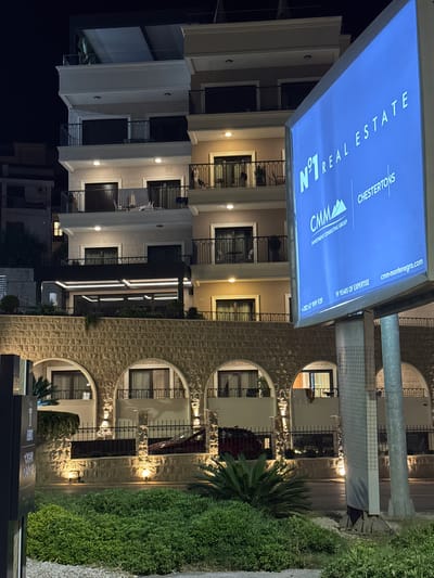 A night-time, low-angle shot captures the facade of a modern building with multiple balconies and arches, illuminated by warm outdoor lighting. In the foreground, lush green bushes and manicured plants are visible, along with a dark sign on the left with some illegible text. Dominating the right side of the frame is a large, illuminated blue billboard displaying advertising text for "N°1 REAL ESTATE" and "CHESTERTONS". The building features a stone facade with arched entrances on the lower level and lighter-colored walls with numerous windows and balconies on the upper floors. The overall impression is one of a well-lit, upscale commercial and residential property in a developed area.