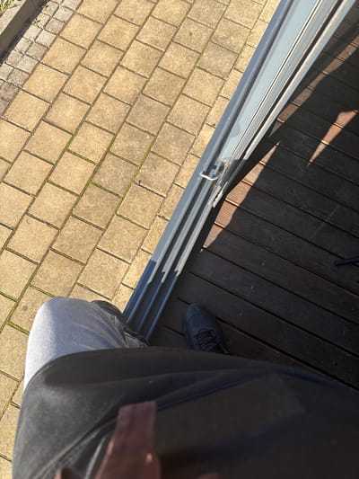 The image depicts a high-angle, first-person view of a person's lower body standing at the threshold of a building entrance or exit in Lom, Bulgaria. To the left, an outdoor area is paved with light tan square concrete blocks, arranged diagonally, with visible moss or weeds growing in the grout lines. A concrete curb runs along the far left edge of the paved section.
A grey, multi-track metal frame, consistent with a sliding door or window system, diagonally bisects the upper middle of the frame, separating the paved area from a dark brown wooden deck on the right. A small, white hook-and-loop mechanism is attached to the lower section of this frame.
The right side of the image features a dark brown wooden deck composed of horizontal planks, some of which are illuminated by direct sunlight, casting distinct shadow lines.
In the lower portion of the image, the person's body is visible, looking downward. The left leg is clad in light blue-grey denim jeans. The right foot, positioned on the wooden deck, wears a dark grey or black athletic shoe with laces. A large, dark object, possibly a bag or garment, is partially visible across the person's lap, with a small brown strap detail at the very bottom edge of the frame. The scene is brightly lit by natural sunlight, indicating an outdoor daytime setting.