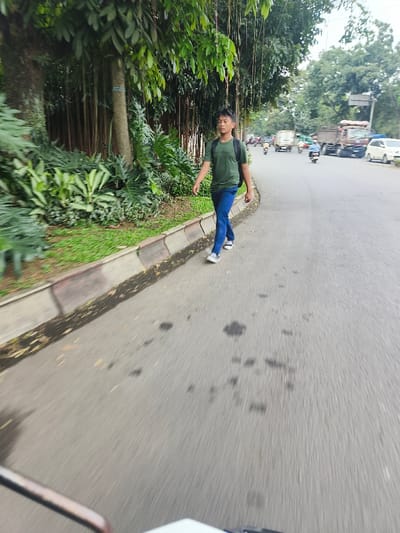The image captures an outdoor street scene in Cimahi, Indonesia, from a low-angle perspective, likely from a moving vehicle. A young male is depicted walking on a concrete sidewalk along the left side of the frame. He is dressed in a dark green short-sleeved t-shirt, blue jeans, and white low-top sneakers, carrying a dark backpack on his shoulders. His dark hair is styled short, and he is looking forward with a neutral expression.

To the left of the male, a dense strip of green vegetation runs parallel to the sidewalk, consisting of various trees with prominent trunks, numerous hanging aerial roots, and large-leafed plants and shrubs. The sidewalk is bordered by a raised concrete curb. To the right of the male, an asphalt road extends into the distance, exhibiting wear, dark stains, and significant motion blur. Multiple vehicles are visible on the road in the background, including several motorcycles, a light-colored car, and a large brown truck, suggesting active traffic. The sky overhead appears overcast. A portion of a white vehicle, possibly a scooter or motorcycle, is visible in the bottom left foreground, consistent with the camera's low and dynamic position.
