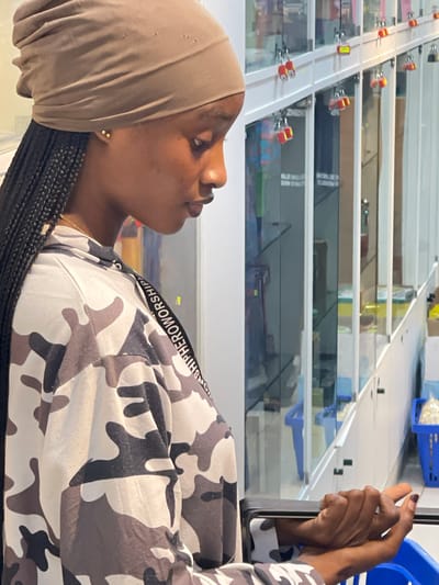 The image shows a young Black woman wearing a beige headscarf and a camouflage-patterned shirt. She is looking upwards and to her right, with her hands near her face. Her right hand is raised, and she appears to be holding a phone, looking at its screen. Her left hand is resting on her chest. She is wearing small gold earrings. Her hair is braided. The background appears to be an indoor setting, possibly a market or a warehouse, with multiple shelves or display cases visible. The shelves are transparent, allowing a view of items stacked behind them. Some of these items appear to be in blue bins. A conveyor belt system with red and yellow components is visible in the upper left portion of the image, suggesting a retail or distribution environment. The lighting suggests it is daytime, but the exact time is not discernible. The overall impression is of a casual moment captured indoors. The text "WORSHIPHERWORSHIP" is visible on a band around her shirt. The location context provided is Guzape, Nigeria.