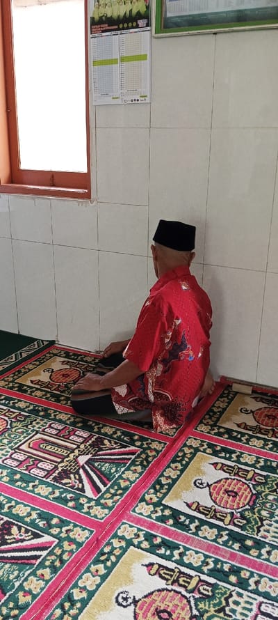 A man wearing a red and black patterned shirt and black hat is kneeling on a prayer rug. He is facing a white tiled wall with a window in the background. The window is covered by a brown frame and is mostly obscured by sunlight.  A calendar hangs on the wall. There is another prayer rug partially visible in the foreground. The scene is set inside a room in a building located in Kecamatan Kutawaringin, Indonesia.