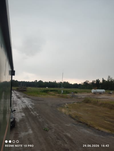 This image captures an outdoor scene on an overcast day, likely in the late afternoon given the timestamp. The foreground features a wet, muddy dirt road, suggesting recent rain or a damp environment. To the left, the side of a large, dark green structure, possibly a building or vehicle, is visible, reflecting the dull sky. A black car is parked on the side of the road.
The midground opens up to a grassy field bordered by a dense line of trees. Scattered within the field are industrial elements, including a large white cylindrical tank with a yellow platform on top, and another smaller white tank in the distance. A tall metal pole with some equipment attached stands near the tree line. Piles of wooden logs are stacked on a wooden frame in the field.
The sky is heavy with clouds, creating a subdued and muted light. The overall atmosphere is one of a working or industrial area in a rural or semi-rural setting. The timestamp "29.06.2026 16:42" indicates the date and time the photo was taken, suggesting a future date. The text "REDMI NOTE 10 PRO" is also visible in the bottom left corner, identifying the device used for capture. The location context provided is Ровенскаслабодскі сельскі Савет, Belarus. There are no visible people or strong emotional cues.