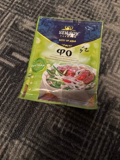 A packet of Sen Soy Pho broth base mix, a product of Asia, is lying on a dark gray carpet. The packet is predominantly light green with a dark blue top section featuring the brand logo and text. The front of the packet displays an image of a bowl of Vietnamese Pho soup, complete with noodles, meat, and herbs. There are also some handwritten markings in what appears to be black ink on the packet. The overall setting is domestic, with no clear indication of a specific city or location, including Chernogorsk, Russia.