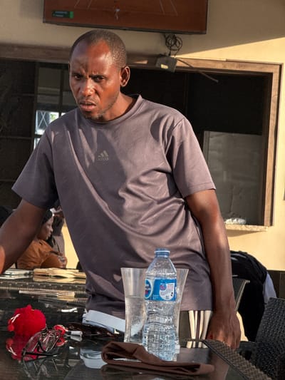 A man in a gray Adidas shirt is seated at a table in what appears to be an outdoor or semi-outdoor setting. On the table are a bottle of water, a glass, sunglasses hanging from a red fuzzy keychain, and a brown garment or cloth. The background includes a window and a building facade. The man looks directly at the camera. The city and specific location within Rubuchi, Nigeria, cannot be determined from the image.