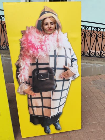 A woman's face is inserted into a cutout of a cardboard standee displaying a person in a hat, a pink feather boa, and a checkered coat. The standee is yellow and appears to be on a sidewalk. In the background, there is a black metal fence and a wall. The ground is paved with bricks. The scene appears to be a photo opportunity setup. The location is identified as Yaroslavl, Russia.