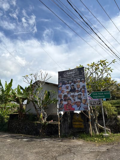This image depicts a roadside scene in Penebel, Indonesia, under a cloudy sky. A large banner displaying a group of men wearing traditional white headwear and clothing is prominently featured. The banner has text in Indonesian that appears to be a greeting or announcement. To the right of the banner, two green directional signs are visible. The upper sign reads "Puluk Puluk Kelod" and "Tingkihkerek," and the lower sign indicates "KAMPOENG JALAK BALI" with a distance of "1.5 km." In the background, a modest building and lush green foliage, including banana trees, are present. Power lines stretch across the top of the frame. The ground is paved, and some scattered yellow flowers are visible near the base of the signs. The overall impression is a snapshot of a local community event or commemoration in a rural Indonesian setting.