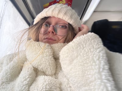 A close-up, chest-up image depicts an individual, appearing female, with light-colored hair and fair skin, looking directly at the camera with a neutral expression. She is wearing round, thin-framed glasses, a cream-colored knitted beanie, and a cream-colored, textured, fluffy jacket or blanket. Her left hand is partially visible, positioned near her temple, touching the beanie or hair. The background indicates an indoor environment, possibly a moving vehicle, with a window on the left showing streaks of condensation or precipitation. A dark-colored object, potentially a seat, is visible in the upper right background, and a small, colorful detail with yellow, red, and blue elements is present above the window on the right. The specific location within the city of Борисово, Russia, cannot be ascertained from the visual evidence provided.