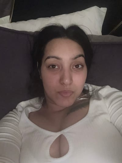 A female individual is depicted in a close-up, head-and-torso selfie, reclining in an indoor, dimly lit environment. She possesses dark hair, brown eyes, and light-to-medium skin. Her face is directed towards the camera with a neutral expression and slightly pursed lips. She is wearing a light-colored, long-sleeved top, possibly white or off-white, featuring a ribbed or textured fabric and a keyhole cutout above the cleavage. The individual is positioned against a dark-colored upholstered surface, likely a sofa or bed. A white pillow is partially visible behind her head, to the upper left of the frame. No discernible features within the image indicate a specific location in Kempten (Allgäu), Germany. The scene is static, with no evident interactions.