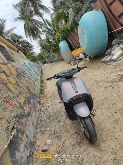 The image shows a grey and black electric scooter parked on a sandy path in Phan Thiết, Vietnam. To the left of the path is a concrete wall covered in graffiti, and beyond that, a building with a window. To the right of the path, there are several large, blue, boat-like objects, some yellow plastic items, and piles of broken concrete. Palm trees line the area, and the sky is overcast. The scooter has a "GO" emblem on the front and "MOTOR THAI" written on the handlebar. The timestamp "2026 07 06 11:09" and the text "realme Shot by dog.lover" are overlaid on the bottom right corner of the image.