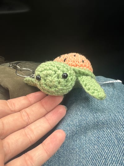 A close-up, eye-level shot shows a crochet turtle keychain. The turtle is predominantly light green with a coral-colored shell speckled with black dots. It has two black bead eyes and a small green flipper visible. A metal keychain ring and clasp are attached to the turtle's head. The turtle rests on a textured blue fabric, likely denim, which is slightly wrinkled. In the foreground, a person's hand, with visible lines and skin texture, is positioned to the left of the turtle, its fingertips gently touching the denim. The background is dark and out of focus. This image was taken in Wattrelos, France.