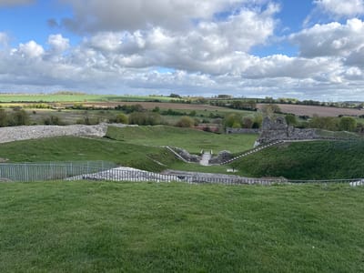 This panoramic image presents a historical site nestled within a vast, undulating rural landscape under a dynamic sky. In the immediate foreground, a well-maintained grassy slope leads down to a dark grey metal railing that curves across the frame, partially obscuring a lower section of the site.
The central feature is a large, semi-circular or amphitheater-like depression carved into the landscape. Its steep, grassy sides are punctuated by remnants of stone walls and multiple sets of stone steps descending into the basin. A light-colored wooden bridge spans a lower area within the depression, connecting different sections of the ruins and pathways. Additional metal railings are visible along the pathways, hinting at visitor access.
Beyond this ancient structure, the background unfolds into a expansive countryside. A mosaic of agricultural fields, ranging in shades from vibrant green to earthy brown, stretches towards the horizon. Clumps of trees and hedgerows are scattered across the rolling hills. The sky is a prominent feature, adorned with large, fluffy white and grey cumulus clouds interspersed with patches of bright blue, indicating a day with mixed sun and cloud cover.
The scene is devoid of people, conveying a sense of tranquility and historical depth. The natural daylight suggests it is either late morning or early afternoon. The combined elements of ancient stone ruins, prominent earthworks, and the surrounding agricultural land strongly indicate a significant historical site, characteristic of locations like Castle Acre Castle in King's Lynn and West Norfolk, United Kingdom. No visible text is present in the image.