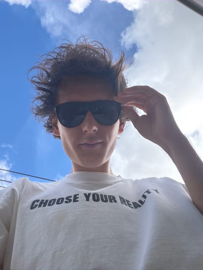 A low-angle, medium shot captures a person from the chest up, against a bright blue sky with scattered white clouds. The individual is wearing a white t-shirt with the black text "CHOOSE YOUR REALITY" printed across the chest. They have dark, tousled hair and are wearing black sunglasses, with their right hand adjusted on the sunglasses' frame. The person's face is turned slightly to their left. The background shows power lines in the upper left corner. The scene appears to be outdoors in the city of Si Sunthon, Thailand, under a sunny sky.