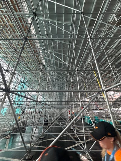 The image shows a complex network of metal scaffolding inside a large structure. The scaffolding is predominantly silver in color, creating a dense, interconnected grid. The ceiling above the scaffolding is corrugated metal. In the upper right corner of the image, there are glimpses of people wearing black hats and orange shirts. The overall environment appears to be an indoor construction or renovation site. The location is most likely in Miami Gardens, Florida, United States of America.