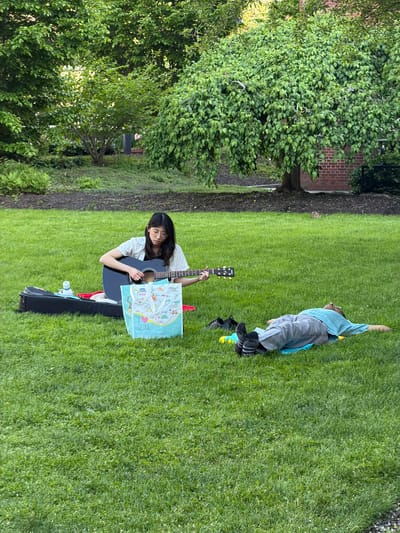 A woman sits on the grass in what appears to be a park or yard, holding a guitar and a shopping bag that reads "I <3 SEOUL." Next to her is a guitar case and a water bottle. A young person lies in the grass nearby with one arm raised. A tree stands to the left of the woman, and a brick building can be seen in the background. Given the possible clues in the description of the location, it is possible to assume this image was taken in Cambridge, Massachusetts, United States of America.