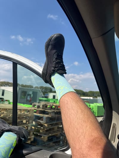 A person's leg and foot are extending out the open window of a vehicle. The person is wearing a light blue sock with a green pattern and a black running shoe. Outside the window, a large quantity of wooden pallets, some with green paint, are visible. The sky is blue with scattered clouds. The location is Mid Suffolk, United Kingdom.