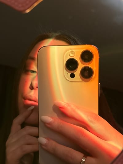 A woman holds an iPhone in front of a mirror, capturing a reflection of her face. The phone's camera lenses are visible, and a beam of warm light illuminates the phone and the woman's hand, accentuating her light-colored nails and a ring on her finger. The overall scene creates a warm, intimate atmosphere.