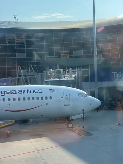 The image shows a Malaysia Airlines passenger airplane at an airport gate. The plane's nose is visible, with the airline's name and the Malaysian flag on its side. The airplane is parked at the airport terminal, which has large windows. A jet bridge is connected to the plane. The ground outside the terminal is visible. The location of the image is likely Kuala Lumpur International Airport (KLIA) in Sepang, Malaysia.