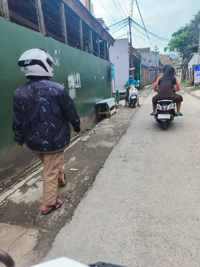 A man wearing a white helmet and a camouflage jacket walks down a street in Cimenyan, Indonesia. He is walking on a paved road with a green wall to his left. There is a woman riding a motorcycle in front of him. The motorcycle is a white Honda Beat with the license plate D 4728 JH. The woman is wearing a brown dress. The man is facing away from the camera and walking towards the woman. In the distance there is another woman riding a white motorcycle. The road is surrounded by houses and trees. There is a sign in the distance that says "ATM."