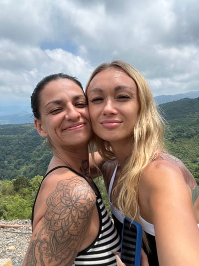 Two women are taking a selfie outdoors. The woman on the left has dark hair pulled back and is wearing a black and white striped tank top. She has extensive arm and shoulder tattoos, including a large mandala design. The woman on the right has blonde hair and is wearing a white tank top. She has tattoos on her shoulder and upper arm. They are positioned in front of a lush, mountainous landscape. The sky is cloudy with patches of blue. The foreground shows a rocky ground. The location is described as Nam Hải, Vietnam.