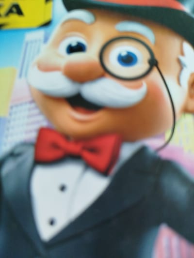 The image is a close-up, blurred photograph of a cartoon character, appearing to be Mr. Monopoly, or a similar figure. He is wearing a black top hat with a red band, a monocle over his left eye, a white mustache and eyebrows, a red bowtie, and a black tuxedo jacket over a white shirt with two black buttons. The background is out of focus and appears to be a cityscape with buildings. The image does not provide any information about the location in the city of Крывасельскі сельскі Савет, Belarus, as the subject matter is entirely artificial and does not depict any real-world location.