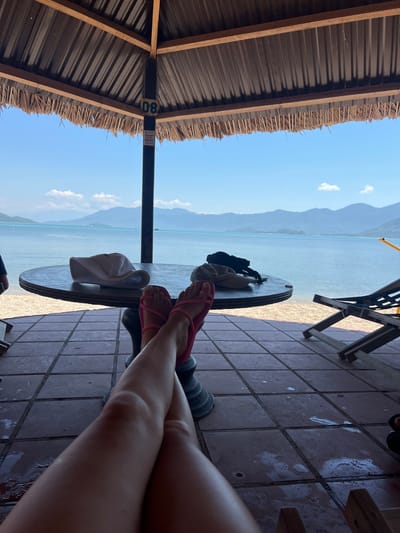 Under a thatched roof shelter, a person's legs are crossed in the foreground, wearing pink flip-flops. A round table with a white hat and two black items sits in front of them. Beyond the table, a tranquil turquoise sea stretches to the horizon, with distant mountains under a bright blue sky. A wooden lounge chair is visible to the right. The scene appears to be a beachside cafe or resting area in Ninh Ích, Vietnam, given the geographical context.