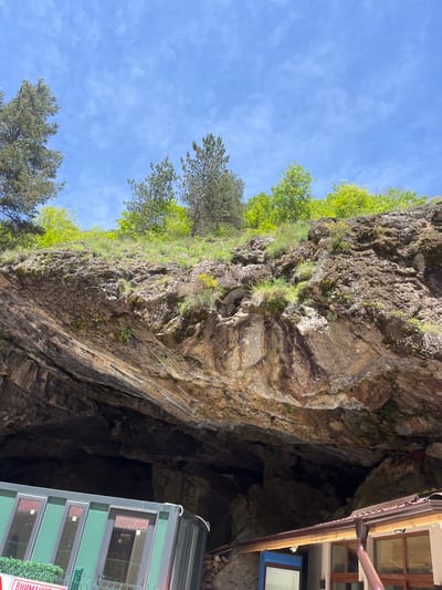 The image depicts a rock formation, possibly a cave entrance, located in Devin, Bulgaria. The rock face is covered in vegetation, including trees and shrubs. To the right, a building with windows is partially visible. The sky is blue with scattered clouds. A green structure with windows, potentially a modern addition to the site, is situated adjacent to the rock face. The entrance to the cave appears dark and cavernous.