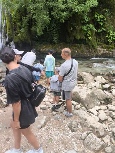 A group of people are gathered near a rocky riverbank at the base of a waterfall in Pirveli Balda, Georgia. The waterfall cascades down a moss-covered cliff face, surrounded by lush green vegetation. Several individuals, including adults and children, are visible in the foreground and middle ground, some standing and others in motion. They are dressed in casual attire. The river water appears clear with visible rocks and currents. The ground is covered in a mix of pebbles, rocks, and some dirt.