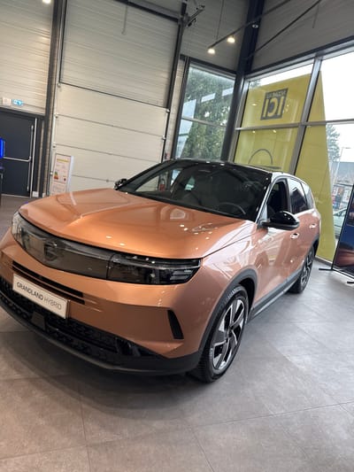 A metallic copper-rose gold Opel Grandland Hybrid SUV with a black roof and trim is displayed inside a car dealership showroom in Hoenheim, France. The vehicle, featuring a distinctive front fascia with integrated headlights and a "GRANDLAND HYBRID" badge on its lower grille, rests on a light gray tiled floor. The showroom environment includes a high ceiling with exposed metal beams and track lighting. To the left, a dark gray exit door and a large white sectional overhead door are visible, along with a yellow informational poster. To the right, large windows offer a view of trees and an external building. A prominent yellow sign, visible through or reflected in the windows, displays the mirrored French text "Nouveau ICI." No individuals are present within the scene, and the vehicle is stationary.