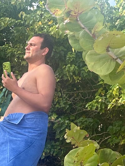 A man wearing a blue towel and holding a green smartphone is standing in a tropical outdoor setting with lush green foliage and large, round leaves overhead. The location is potentially near a beach in Sainte-Anne, France, based on the tropical vegetation. The man is looking at his phone, and the lighting suggests it's daytime. The environment is full of dense greenery, indicating a natural or semi-natural area.