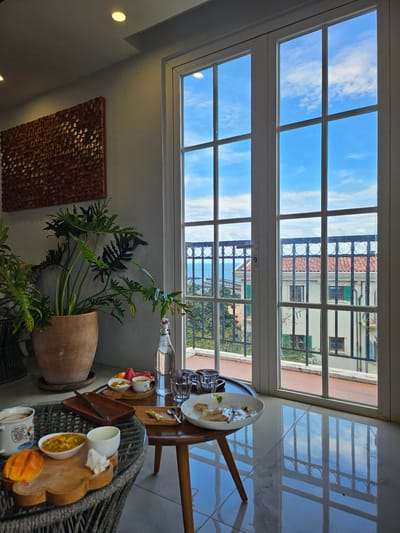 The image shows a still life scene in an interior space, likely a cafe or restaurant, with a view of a balcony and buildings beyond. In the foreground, two tables are visible. The table on the left is round and made of rattan, featuring a white mug with a cartoon print, a white bowl containing passionfruit, a white ceramic cup, and a wooden board with sliced mango and what appears to be a piece of white napkin. The table on the right is a darker wood with a rectangular wooden tray holding a small cup and some fruit slices, a small white bowl, a glass of water, and a white plate with remnants of food and a fork. A bottle of water stands behind this table. A large potted plant with lush green leaves is situated to the left of the tables. The background features a large window with white frames, divided into multiple panes, looking out onto a balcony with a black wrought-iron railing and red-tiled roofs of buildings. The sky outside is blue with white clouds, reflecting on the polished floor. The overall impression is of a relaxed setting with a meal or snack in progress, overlooking a picturesque view. The prompt mentions Phu Quoc, Vietnam, suggesting this scene is located there.