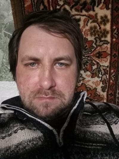 A mid-40s Caucasian man is looking directly at the camera. He has short, dark brown hair and a beard. He is wearing a black and white striped knit sweater. Behind him is a rug with an ornate brown, red, and green floral pattern. To his left is a pale green wall with a white painted texture. The location of the scene is Parabel, Russia.