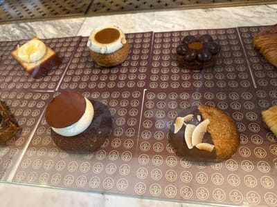 An assortment of pastries is displayed on a marble countertop behind a glass barrier, likely in a bakery or patisserie in Copenhagen, Denmark. From left to right, the visible pastries include a square cake topped with white frosting and a yellow garnish, a round pastry with a light brown bun, white frosting, and a caramel filling, a dark chocolate pastry shaped like a flower with a small orange center, and a golden croissant. Below these, another row of pastries is visible: a slice of dark cake, a round chocolate pastry with white frosting and cocoa powder dusting, and a pastry coated in coconut flakes with additional coconut pieces on top. The pastries are arranged on individual brown paper liners featuring a repeating white floral or human-like symbol. The background shows a blurred interior of the establishment, possibly with additional display cases or shelving. There are no people visible in the image.