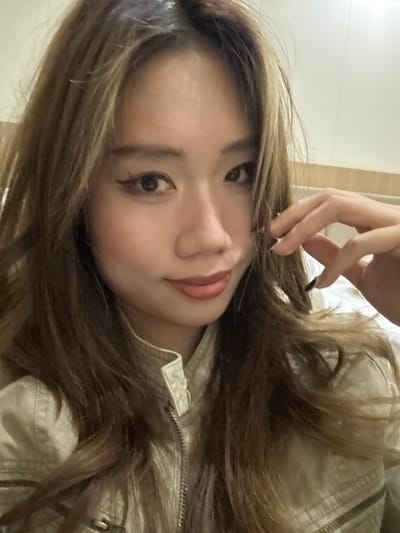 The image captures a close-up selfie of a young woman, with the frame rotated diagonally. She is positioned with her head towards the lower left and her body extending towards the upper right. Her face is directed towards the camera with a calm, slightly alluring expression, her eyes wide open and lips softly parted with pinkish-red lipstick. She has long, light brown hair with noticeable highlights, which appears to be spread around her. Her eyes are accentuated with dark eyeliner and mascara.

She is wearing a light-colored jacket, likely beige or light gray, with a visible zipper running down the front and textured fabric details on the shoulders and collar. One of her hands is gently placed near her temple, touching her hair, revealing fingernails with a distinct design, possibly black tips over a lighter base. The background is indistinct and appears to be a plain, light-colored indoor surface, possibly a wall or ceiling. There are no explicit visual cues within the image to suggest the provided location context of Dunhuang City, China, nor is the time of day or weather discernible. No text is visible in the media file.