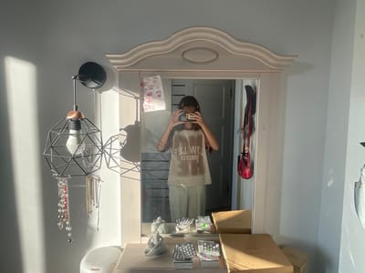 The media file depicts a girl taking a mirror selfie in a bedroom. The girl, wearing a light-colored t-shirt with text and striped pants, is holding a camera up to capture her reflection.

In the background, a mirror is mounted on a light-colored dresser with a decorative, curved frame. The room appears to be well-lit by natural sunlight streaming in from the left, casting distinct shadows on the wall and floor, suggesting it is daytime. Visible objects on the dresser include several small sculptures, blister packs of pills, and cardboard boxes.

To the left of the mirror, a black industrial-style pendant lamp with a metal cage design hangs from the wall. Several necklaces are displayed on the lamp's structure. On the right side of the mirror, a red handbag is hanging. The overall scene suggests a personal space, possibly a bedroom or dressing area. The presence of pills and the casual attire might hint at a private moment. The text on the girl's t-shirt is visible in reverse: "FOREVER LOVE" and "LOW LIFE". The location context provided indicates this is in Мухавецкі сельскі Савет, Belarus.