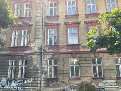 This image depicts a section of an old, weathered building facade in Bielsko-Biała, Poland. The building exhibits signs of age and disrepair, with peeling paint, chipped plaster, and exposed brickwork, particularly on the lower levels. It features multiple stories with a series of tall, rectangular windows, many of which are divided into panes. The window frames appear to be white, and some are surrounded by decorative red trim. The building's exterior is textured, with some areas appearing to be made of large, rectangular stone blocks.
Several trees with vibrant green foliage are visible, partially obscuring parts of the building, with sunlight dappling through the leaves. In the lower portion of the image, graffiti is spray-painted across the wall and some of the windows, adding to the urban and somewhat neglected aesthetic. There are no people visible in the frame. The scene captures a typical urban landscape element, showcasing the architectural style and current condition of a historic building in the city.