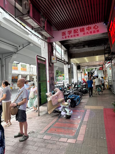 In Yuexiu District, China, a busy sidewalk scene unfolds under an overhead structure. A tall, digital display board, likely for public transport, stands prominently on the right side of the walkway, showing a green interface with rows of text and numbers. Adjacent to it, a column displays various signs, including what appears to be a building or establishment name in red lettering at the top, and a white sign with Chinese characters and a barcode at the bottom.
Several people are present, some waiting near the display board. A man in a striped shirt and dark shorts stands in the foreground, his gaze directed forward. To his left, a woman in a striped shirt and light-colored pants waits patiently. Further back, a woman in a blue top and a woman in a floral skirt and pink top are visible. A man in a blue t-shirt walks along the sidewalk, and another man in a white t-shirt rides a bicycle, a young child behind him. In the distance, more individuals and a bus are partially visible.
The sidewalk is paved with interlocking bricks. To the left of the display board, a covered area with pillars supports the overhead structure, which is lined with horizontal red slats. The ground in front of the covered area is also paved with bricks, with some metal grates or covers embedded in it. Numerous parked motorcycles and bicycles line the walkway, with some displaying blankets or covers. On the right side of the walkway, a red carpet or mat runs along the edge of a building facade which has several doors and windows, and displays various signs and merchandise. The overall environment suggests a commercial street with a focus on transportation and pedestrian activity.