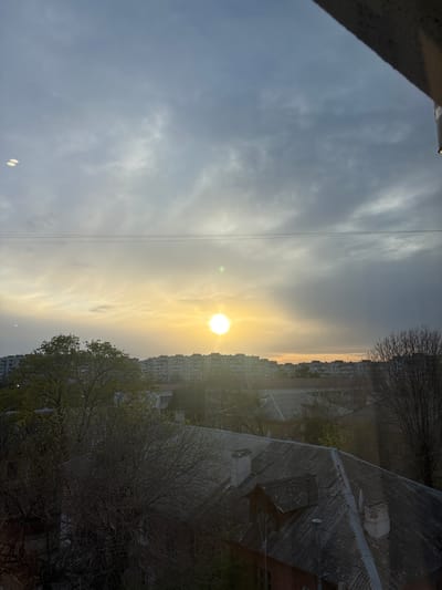 The image captures an urban landscape during late afternoon or early evening, viewed from an elevated perspective, possibly a window or balcony. The bright sun, positioned on the right side of the frame, is low on the horizon, casting a warm, golden glow across the sky and through scattered clouds. Several thin, dark wires or power lines traverse the sky horizontally.
In the mid-ground, a skyline of multi-story residential buildings is visible, some partially silhouetted against the bright sky. The foreground features the dark, textured, corrugated roofs of older structures, including chimneys, a satellite dish, and what appears to be a ventilation pipe. Bare trees with intricate branch patterns, some showing early signs of budding leaves, are interspersed among the buildings, suggesting late winter or early spring. A concrete edge is visible in the upper left corner of the frame. The scene is quiet, with no human activity or visible text. The setting is an urban environment in Simferopol, Ukraine.