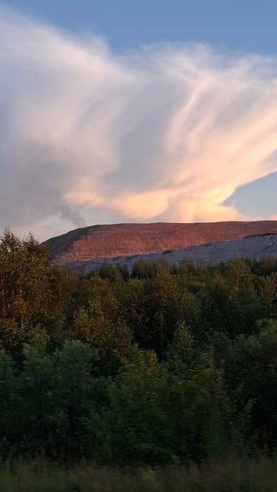 The image shows a vertical rock face with a forest in the foreground. The rock face is illuminated by the setting sun, casting a warm, reddish glow. The sky above is a soft blue with wispy clouds tinged pink and orange. The forest is lush and green, with trees of varying heights. The image appears to have been taken during golden hour, as indicated by the warm lighting and the colors in the sky. The location is Achinsk, Russia, though there are no specific landmarks visible to confirm this. The overall mood of the image is serene and picturesque, evoking a sense of natural beauty and tranquility. No people or text are visible in the image.