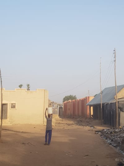 A man balances a white bucket on his head while walking down a dirt street in Dutsinma B, Nigeria. The street is lined with buildings. Power lines extend overhead.
