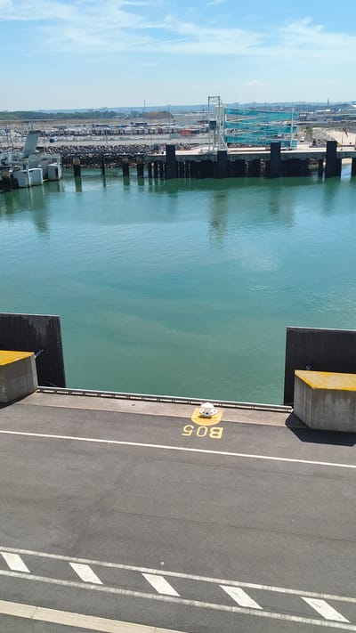 The image captures a view of the Port of Calais in France on a clear, sunny day. The foreground features an asphalt surface with white line markings and a yellow number "B05" painted on the ground, along with a white bollard. To the left and right of the foreground are concrete structures with yellow tops, forming part of the port infrastructure.

The middle ground is dominated by a body of calm, greenish-blue water, likely a part of the harbor. Along the edge of the water are dark, vertical pilings supporting a pier.

In the background, a large ferry terminal is visible, featuring ramps and structures that facilitate vehicle and passenger transfer. Beyond the terminal, a sprawling area with rows of trucks and trailers is spread across the land. In the distance, a low-lying landscape with buildings and some industrial elements can be seen under a bright blue sky with scattered clouds. The overall scene depicts a busy maritime port environment.