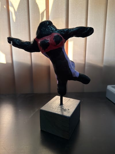 A close-up, low-angle shot shows a black, abstract sculpture of a human torso and limbs, seemingly in a dancing pose. The sculpture is adorned with a red bikini top and purple accents. It is mounted on a thin black rod, which is inserted into a dark grey, cube-shaped base. The base rests on a dark, glossy surface, likely a table. In the background, vertical blinds with a textured pattern create a soft, diffused light, casting shadows onto the wall. A white object, possibly electronic, is partially visible on the right side of the frame. The lighting suggests an indoor setting, potentially a home or studio. This image was taken in Bissendorf, Germany.