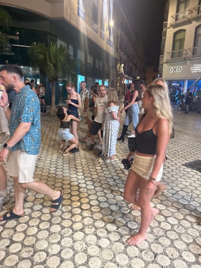 In Málaga, Spain, on a cobbled street at night, a group of people are gathered. A man in a blue patterned shirt and white shorts walks towards the left of the frame. In the center, a woman in a black top and denim shorts is being embraced by another woman. To the right, a woman with blonde hair, wearing a black camisole and a sparkling white skirt, walks away from the camera. Further back, other individuals are seen mingling, with shops and buildings lining the street, some with illuminated signs. The overall scene depicts a lively evening on a pedestrianized urban thoroughfare.