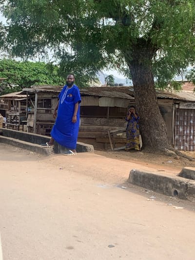 This image depicts an outdoor urban scene in the city of Egbedi, Nigeria. In the foreground, a light brown dirt road spans horizontally, interrupted by two parallel, low concrete structures resembling curbs or drainage channels. The left concrete structure is partially obscured by the subject on the left, while the right structure shows a dark, circular opening on its face.
Two individuals are present. On the left, a dark-skinned adult male with a full beard stands facing the camera. He wears a long, loose-fitting royal blue traditional garment (likely a kaftan or agbada) with white embroidery detailing around the neck and chest. A small, multi-colored emblem, possibly a flag, is visible on his left chest pocket. He wears dark sandals or clogs with prominent white soles; his left foot rests on the far concrete structure, while his right foot is on the dirt road next to it.
To the right, a dark-skinned adult female stands partially concealed behind the trunk of a large tree with a textured bark and visible surface roots. She wears a long, patterned dress, predominantly blue with yellow and black motifs. Her hands are raised to the sides of her head, an action possibly associated with holding a phone or reacting to an auditory stimulus.
Behind the individuals and the tree, a series of rudimentary structures, likely shops or dwellings, are visible. These buildings are constructed from weathered wooden planks and feature corrugated metal roofs, some with evidence of rust. Lush green foliage from various trees forms a dense canopy above the scene and extends into the background, where distant, undulating hills are discernable under an overcast sky.