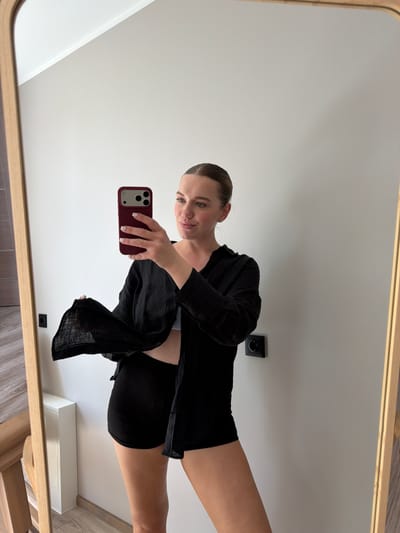 A woman is taking a mirror selfie in what appears to be a well-lit room with white walls and wooden flooring. She is wearing a black open button-up shirt over black shorts. Her hair is pulled back into a ponytail, and she is holding a red-cased smartphone in her right hand, capturing the reflection. Her expression is neutral as she looks at her phone. The background includes a light switch on the wall and a wooden frame around the mirror. The image seems to be taken during daylight hours, given the bright illumination.