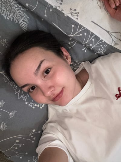 The scene depicts a young adult female positioned horizontally, presumably lying down, within an indoor environment identified as the city of Bayronovka, Russia. The individual has dark hair pulled back from her face, dark eyes, and fair skin, with a faint smile directed towards the camera. She is wearing a light-colored (likely white or off-white) short-sleeved t-shirt featuring a red embroidered design on the right side of her chest, resembling stylized lettering. A small gold-colored hoop earring is visible on her right earlobe, and another small stud earring is present on the upper cartilage of the same ear. She is resting on bedding characterized by a dark grey background with a repeating pattern of white or light-colored botanical elements, including fern fronds and dandelion-like structures. In the upper right corner of the image, a light-skinned hand, possibly belonging to the depicted individual or another person, is partially visible.