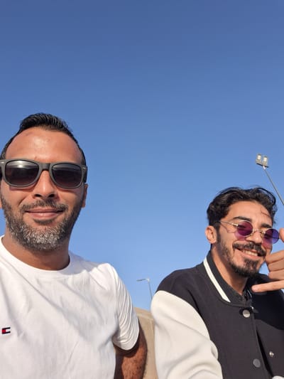 The image captures two adult males outdoors against a clear blue sky in Dar Bouazza, Morocco. The man positioned prominently in the left foreground has dark hair, a beard with visible grey hairs, and wears black square-frame sunglasses, reflecting direct sunlight. He is smiling faintly and wears a white crew-neck t-shirt with a small rectangular red, white, and blue logo on the left chest. A small mark is present on his forehead above his right eye. The man on the right, positioned slightly behind, has dark curly hair, a dark beard and mustache, and wears round, gold-frame sunglasses with purple-tinted lenses. He is smiling broadly, revealing teeth, and is dressed in a black varsity-style jacket with white sleeves and silver snap buttons. His right hand is visible, forming a "shaka" gesture. In the upper right background, a tall pole with two rectangular light fixtures is visible against the uninterrupted blue sky.