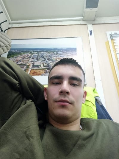 A man in a green sweatshirt poses for a selfie. He has short, dark hair and a neutral expression. Behind him is a framed aerial view of what appears to be an industrial complex. The room has white walls, and a window with horizontal blinds is visible at the bottom. A partial view of a light-colored padded jacket is visible at the top of the frame.